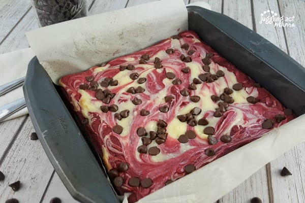 A full pan of red velvet cheesecake brownies with chocolate chips, uncut. It sits on a white wood table with chocolate chips scattered on it.