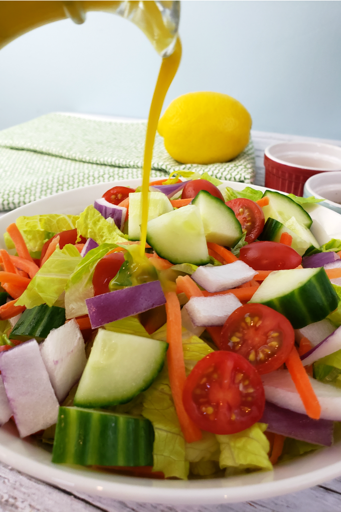 Lemon Vinaigrette Salad Dressing Recipe: Wide bowl full of garden salad with a lemon in the background. A drizzle of healty lemon dressing is poured on the salad.