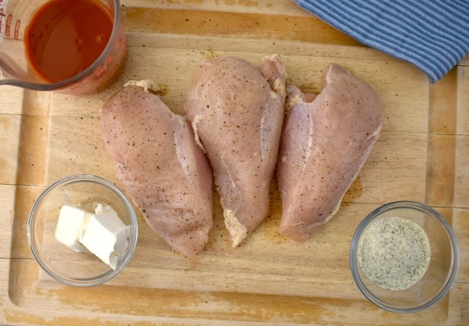 Crockpot Buffalo Chicken ingredients: A cutting board with three large uncooked chicken breasts with a ramekin of melted butter, a mesuring cup with buffalo sauce and ranch seasoning mixture, a ramkein of cream cheese and a blue towel.