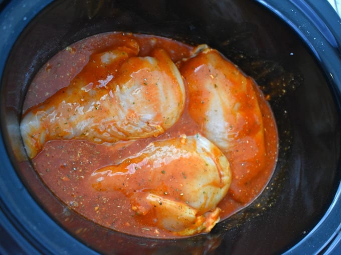 Three large uncooked chicken breasts in a slow cooker crock with buffalo sauce and other ingredients.