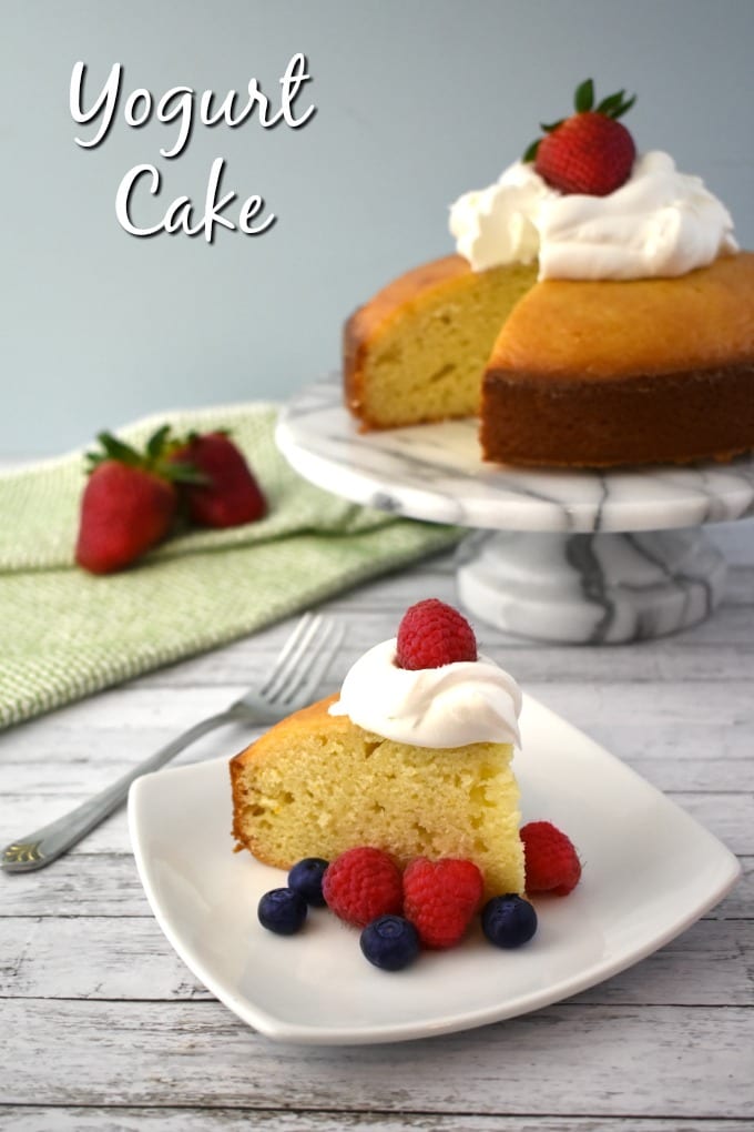 A slice of Yogurt Cake topped with a dollop of whipped cream and surrounded by fresh raspberries and blueberries sits on a white plate. A green and white hand towel can be seen in the background along with a marbled cake tier and a whole Yogurt Cake missing one slice.