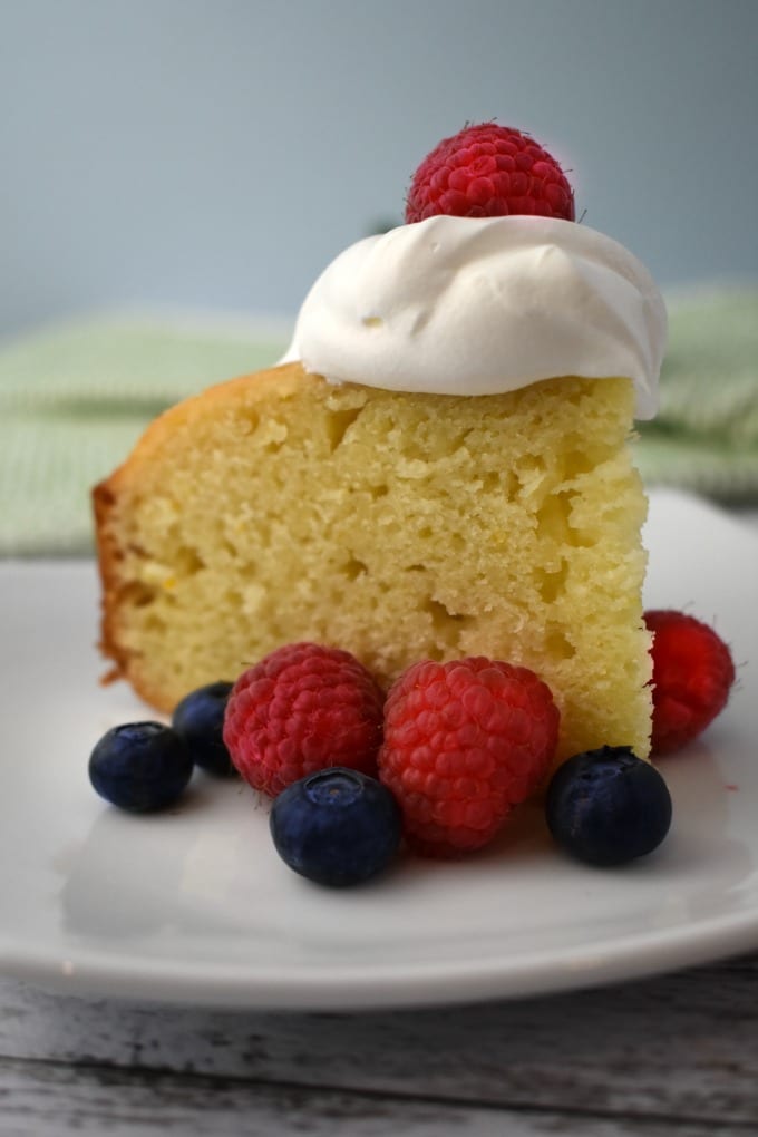 A slice of Yogurt Cake topped with a dollop of whipped cream and surrounded by fresh raspberries and blueberries sits on a white plate. A green and white hand towel can be seen in the background.