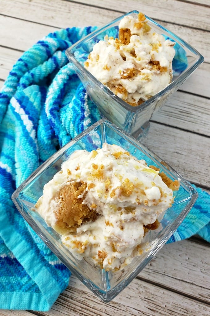 Two stemless martini glasses filled with pineapple delight dessert and garnished with graham cracker crumbs on a wooden surface with a mult-hued blue kitchen towel.