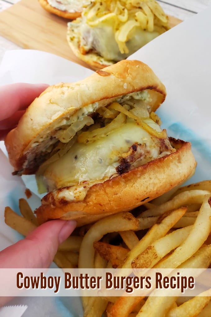 A hand holds a Butter Burger topped with tender onions in a basket with shoe string fries.