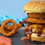 Incredible Cowboy Burger piled high with bacon and crispy onion rings on a sesame seed bun on a slate and wood cutting board. Slices of tomatoes and onion rings can be seen to the left.
