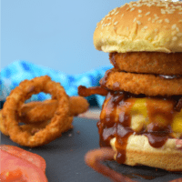 Incredible Cowboy Burger piled high with bacon and crispy onion rings on a sesame seed bun on a slate and wood cutting board. Slices of tomatoes and onion rings can be seen to the left.