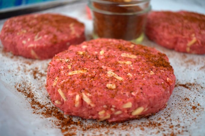 A cowboy burger patty close up showing the cheese mixed in and seasoning sprinkled on top sits on a baking sheet lined with parchment paper. Other patties can be seen in the background along with a jar or cowboy steak rub.