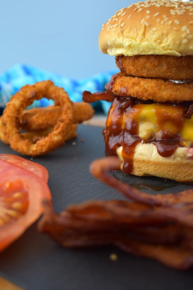 Incredible Cowboy Burger piled high with bacon and crispy onion rings on a sesame seed bun on a slate and wood cutting board. Slices of tomatoes and onion rings can be seen to the left.