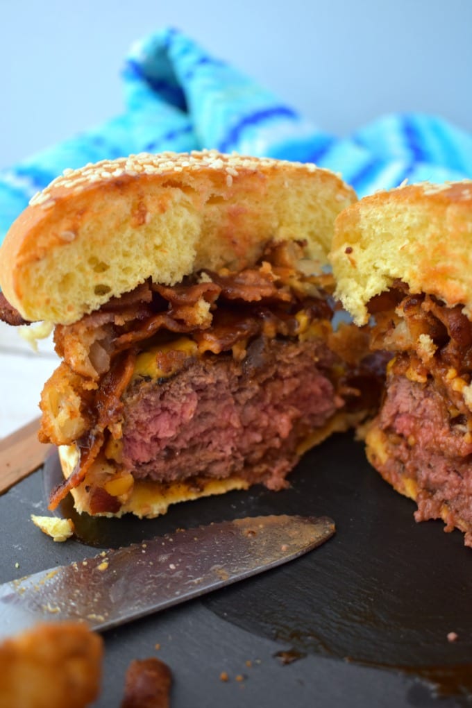 Incredible Cowboy Burger piled high with bacon and crispy onion ringsand sliced in half on a sesame seed bun on a slate and wood cutting board with a knife.