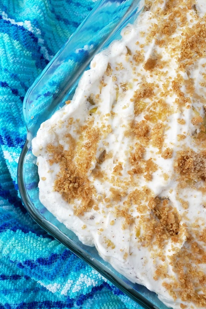 A close up of a baking dish full of pineapple delight dessert sprinkled with graham cracker crumbs on a mult-hued blue kitchen towel.
