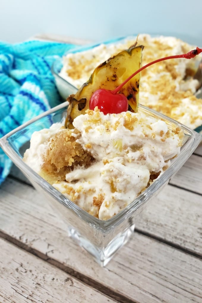A stemless martini glass full of pineapple delight garnished with a cherry and piece of grilled pineapple. In the background there is a square baking dish of the dessert with a multi-hufed blue kitchen towel.