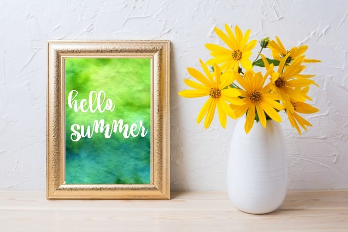 Hello summer free printable displayed in a gold frame next to a white vase with yellow flowers.