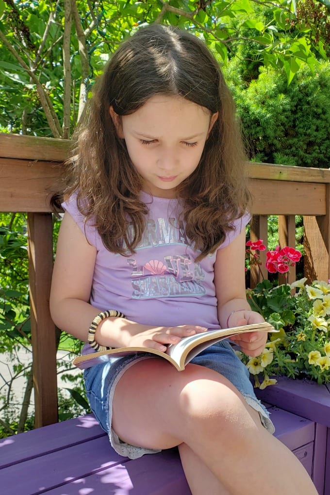 A young girl sits on a a purple Planter Box Bench reading a book.