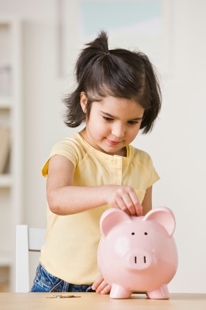 Little girl in a yellow shirt putting coins into a pink piggy bank