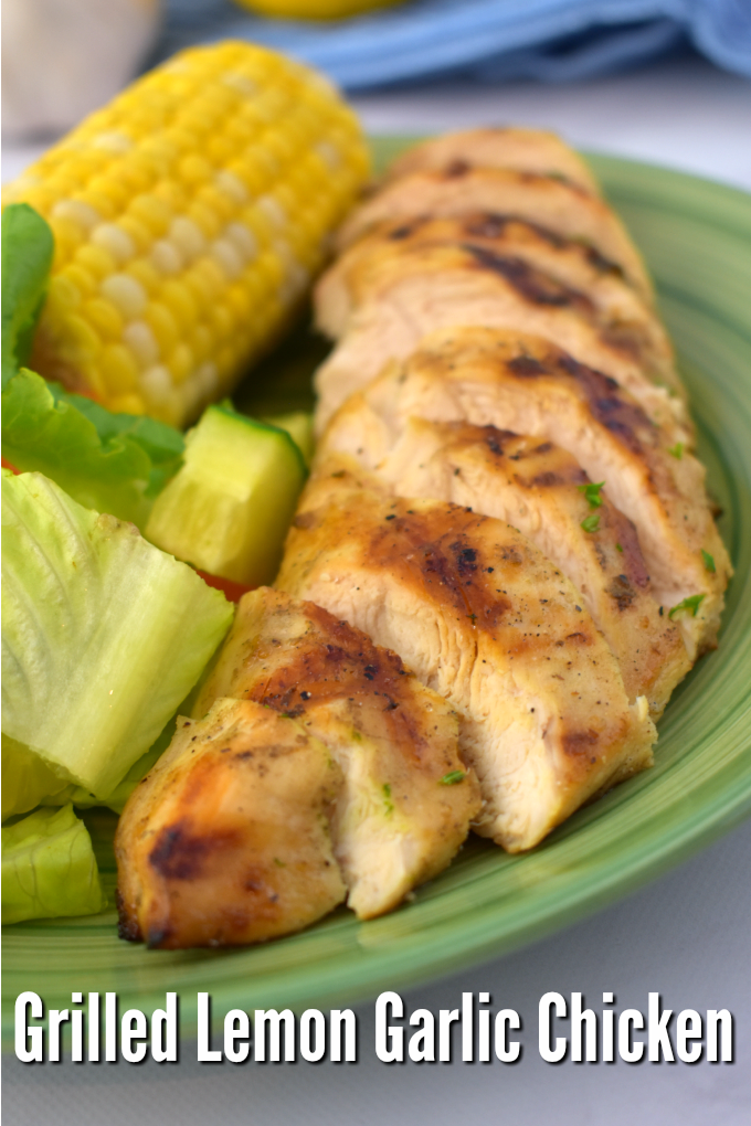 A close up of lemon garlic grilled chicken cut into slices on a green dinner plate next to a garden salad and corn on the cob.
