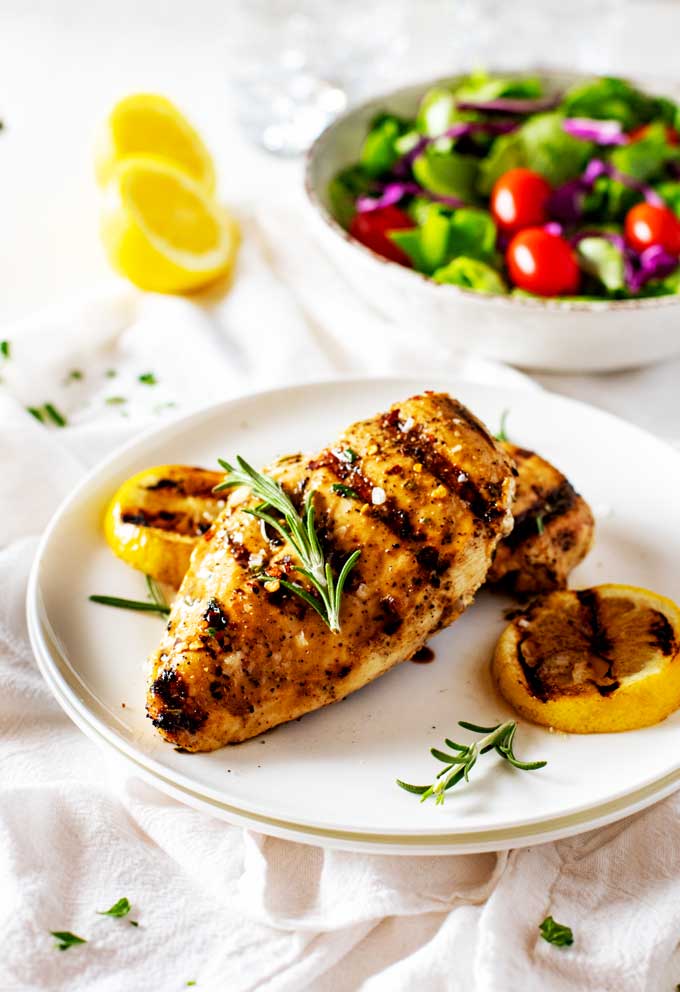 Horizontal image of Grilled Lemon Pepper Chicken on a white plate with a simple salad in a bowl behind it.