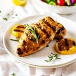 Photo of Grilled Lemon Pepper Chicken on a white plate garnished with rosemary and grilled lemons.