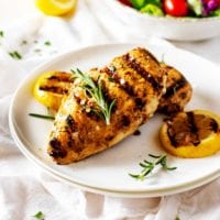 Photo of Grilled Lemon Pepper Chicken on a white plate garnished with rosemary and grilled lemons.