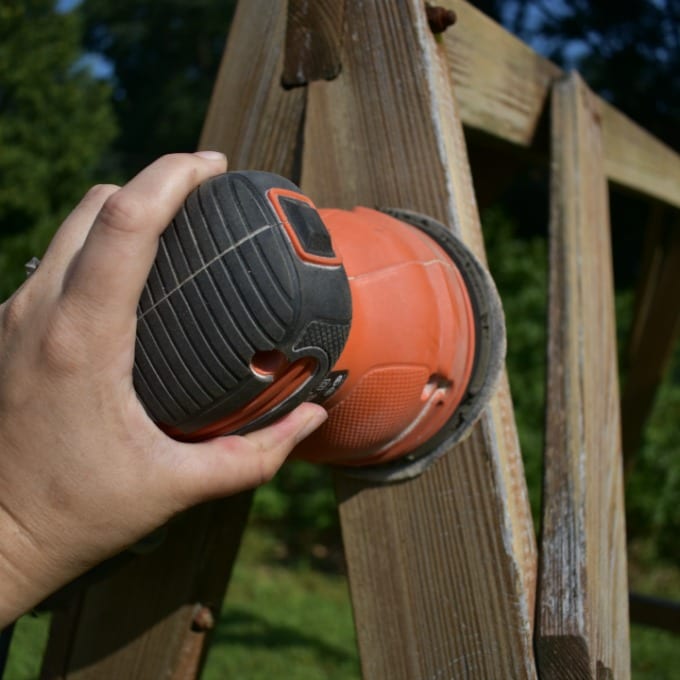 A hand holding an orbital sander working on a shabby outdoor wooden swing.