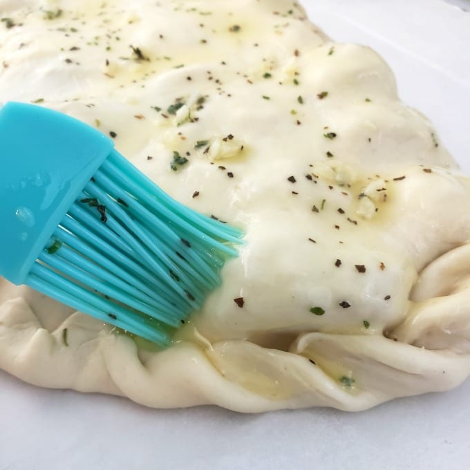 A pastry brush adds melted herbed butter on a calzone dough.