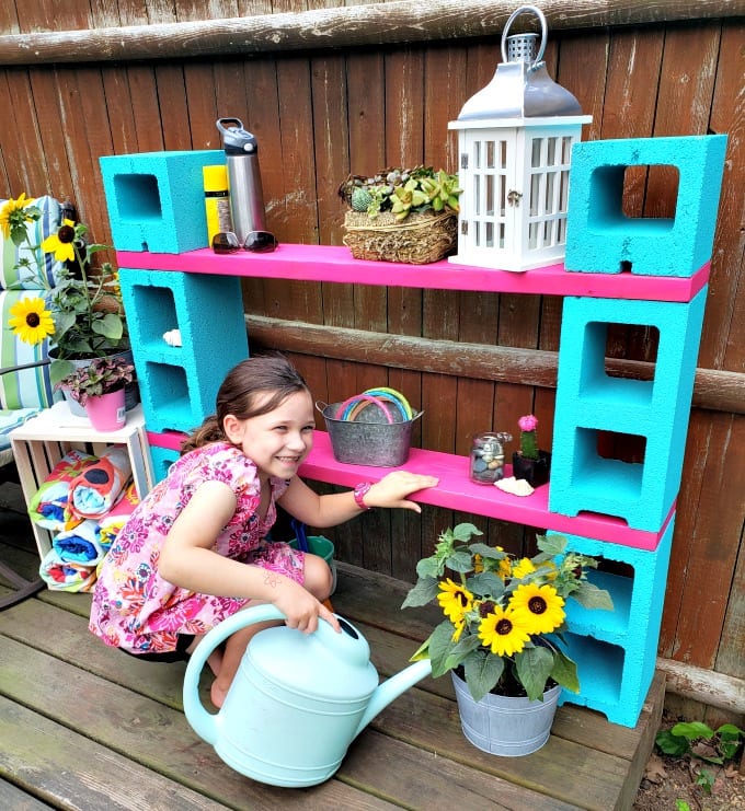A little girl waters a potted sunflower next to a magenta and teal cinder block shelf