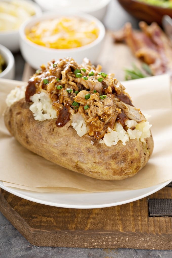 A baked potato on a white plate topped  with pulled pork, BBQ sauce and chives.