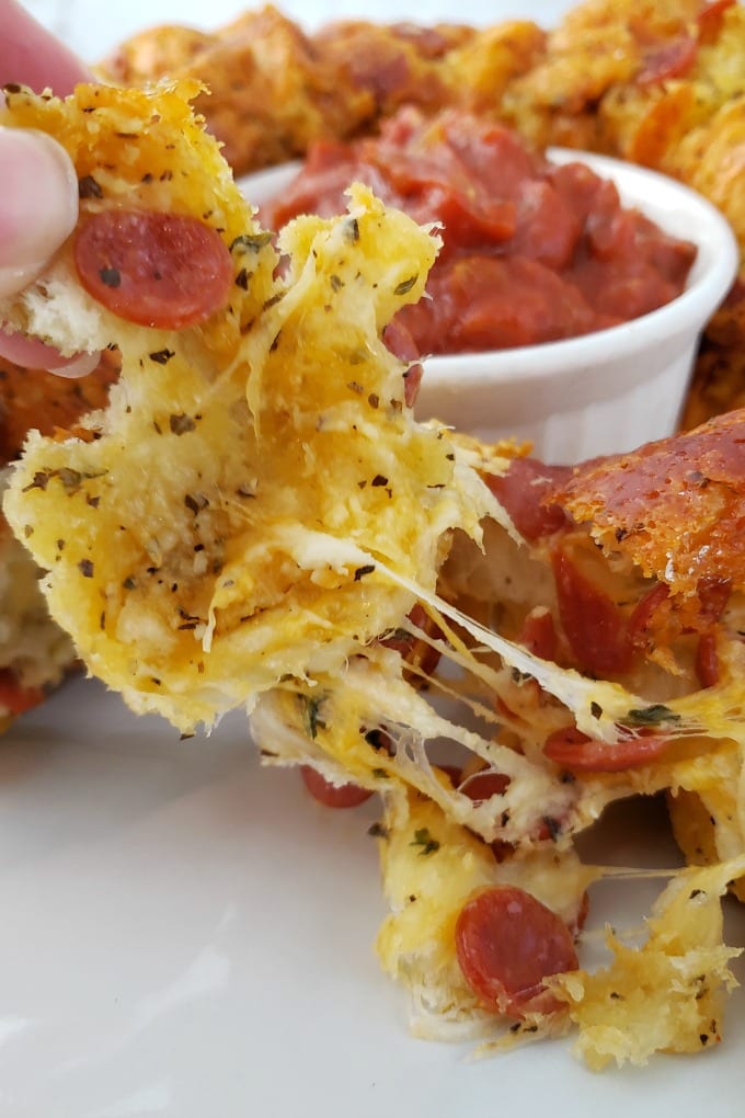 Close up of a hand pulling a piece of cheesy Pull Apart Pizza Bead on white plate next to a ramekin of marinara sauce.