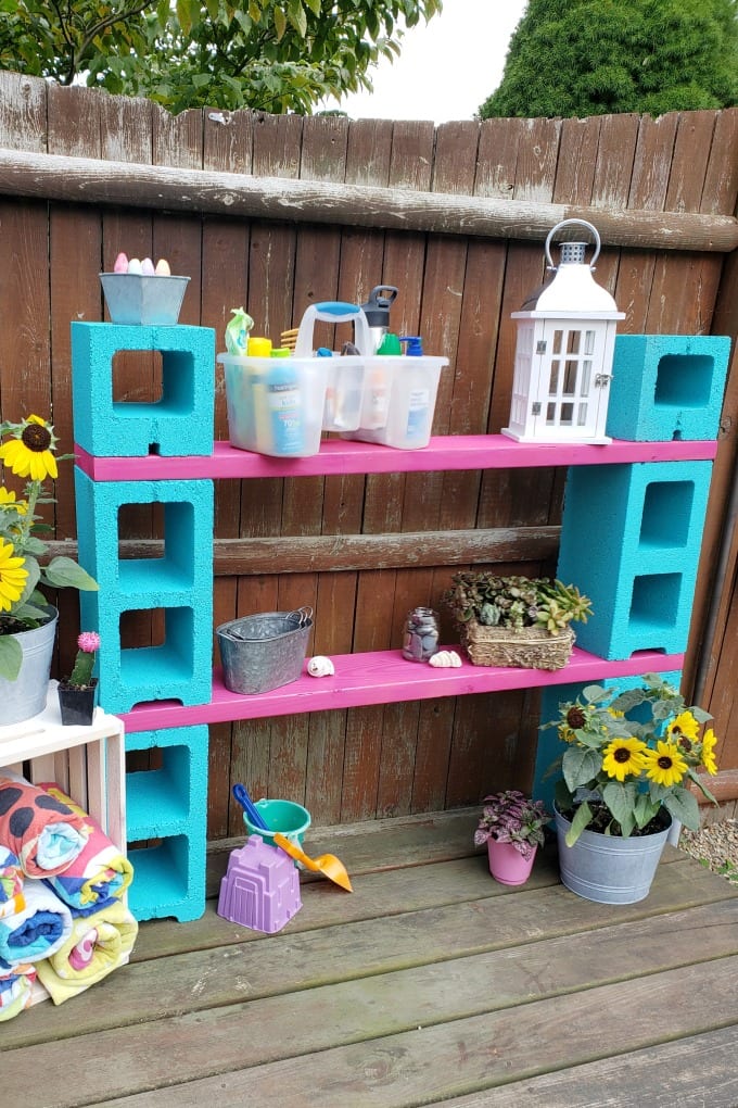 A teal and magenta DIY outdoor storage cinder block shelf next to a fence.