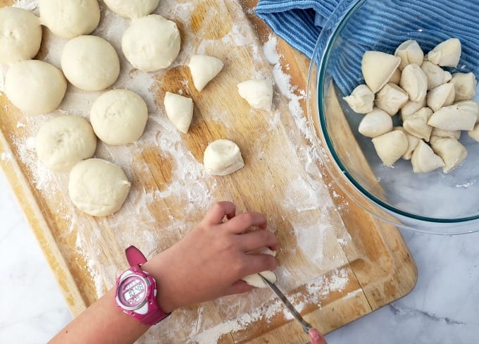Thawed dinner roll dough on a cutting board with a child's hand cutting the dough into quarters.