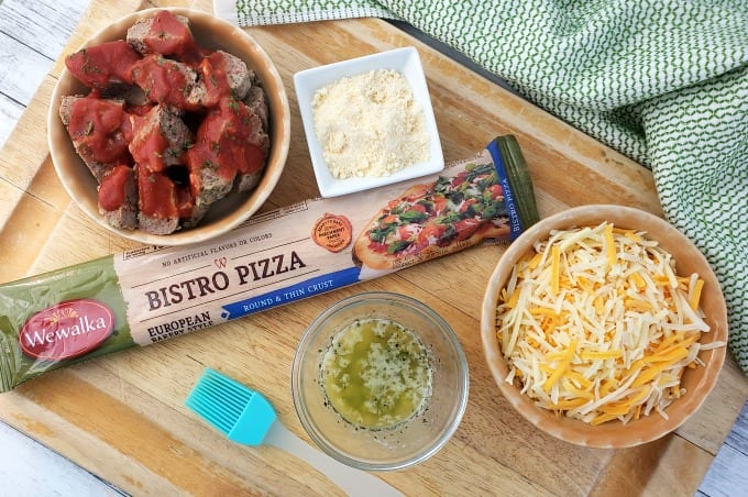 Ingredients to make meatball calzones on a cutting board including Wewalka Bistro Pizza dough, shredded cheese, Parmesan cheese, meatballs with tomato sauce and herbed melted butter.