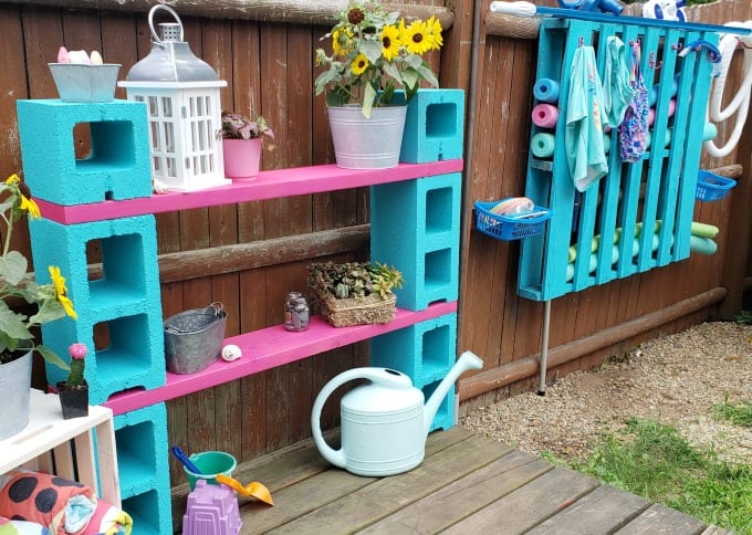 a DIY outdoor storage - cinder block shelf painted magenta and teal in a backyard next to a stained fence.