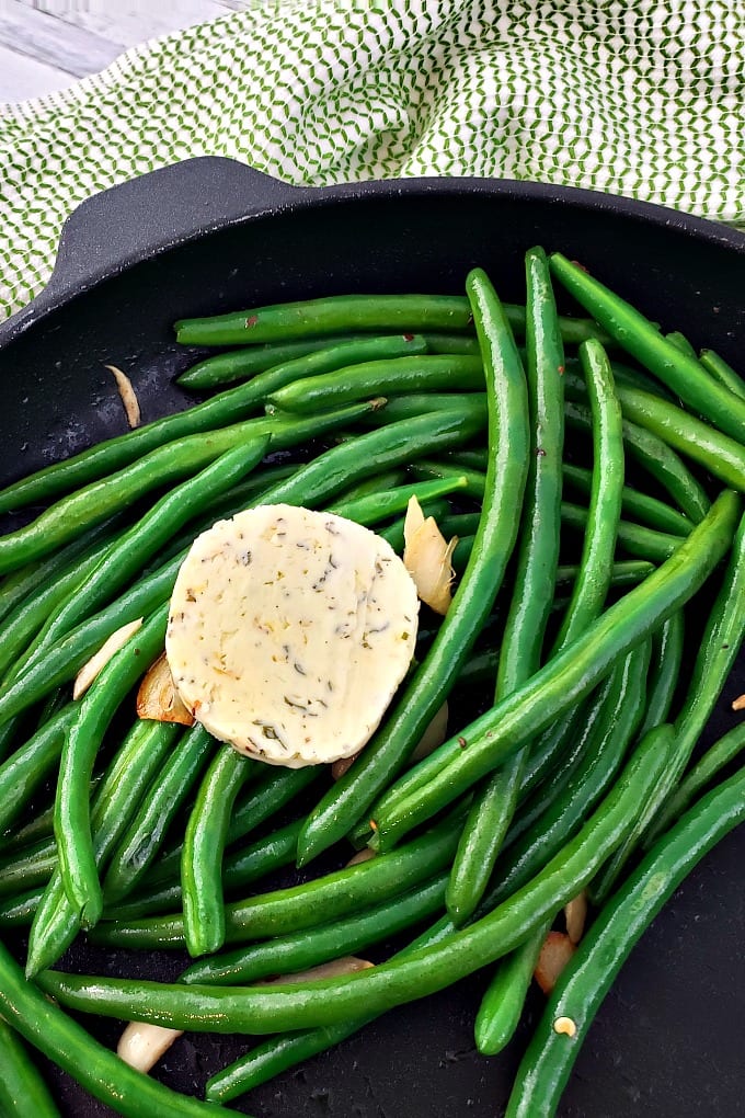 cooked and seasoned green beans in a skillet dish topped with sauteed garlic and compound butter