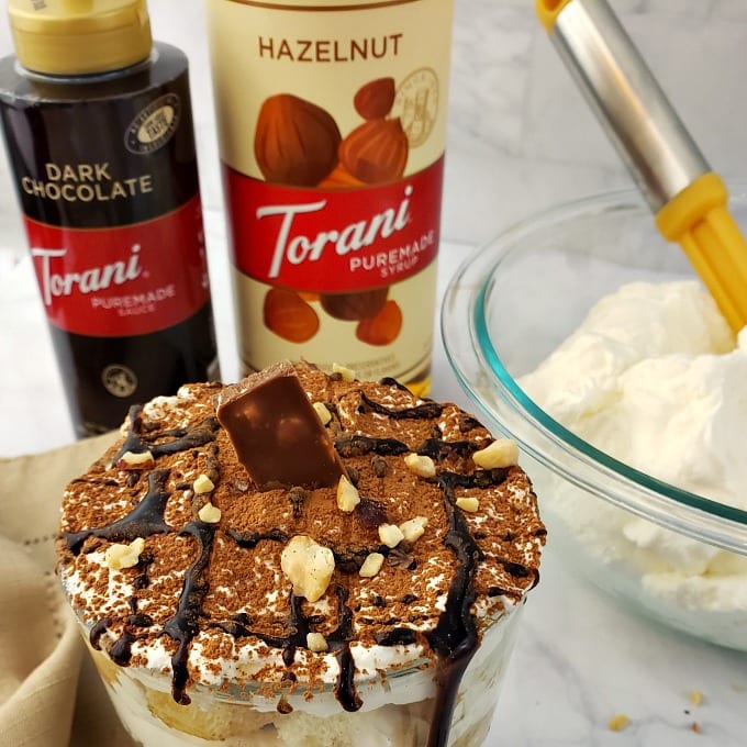 A close up of a Tiramisu Cup dessert drizzled with chocolate and dusted with cocoa powder.
A bottle of Torani Puremade Dark Chocolate Sauce and Torani Puremade Hazelnut Syrup next to a bowl of whipped cream in the background.