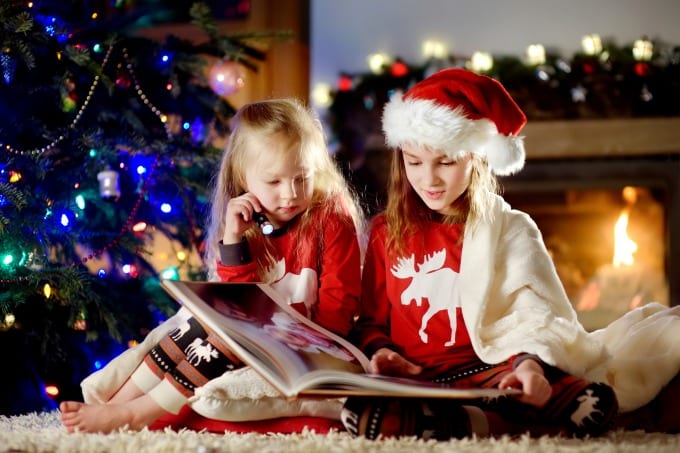 Two girls reading a book in front of a decorated Christmas tree.