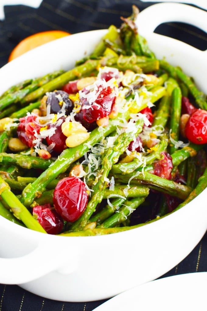A white serving dish of asparagus with toasted pine nuts, fresh cranberries and garnished with Parmesan cheese