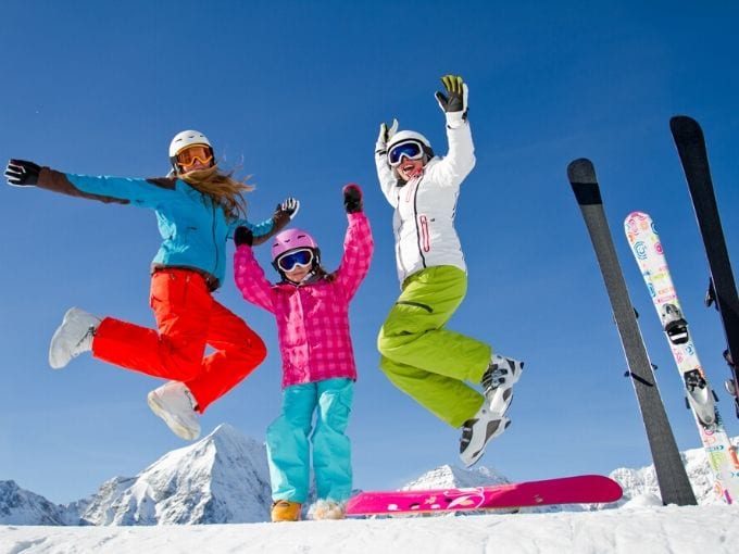 Three girls dressed in bright colored snow pants and ski jackets jumping with lots of joy on their faces