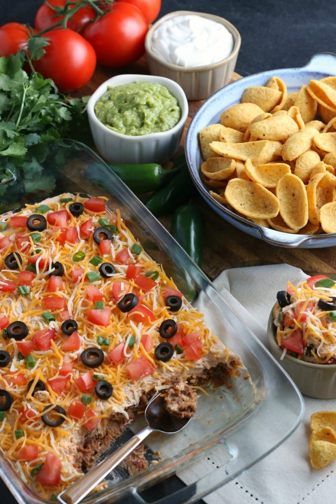 Layered Taco Dip in a clear baking dish next to a bowl of corn chips and other condiments.