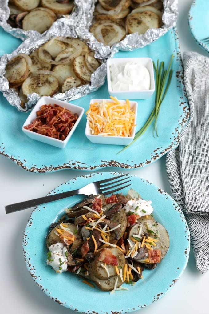 teal plate loaded with best grilled potatoes, yellow onions, and sliced mushrooms topped with bacon, cheese and chives. In the background are dishes of condiments and more foil pouches of potatoes.