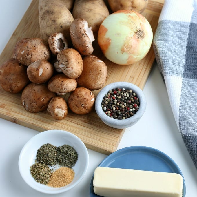 russet potatoes, yellow onion and mushrooms on a cutting board next to a stick of butter on a blue plate and bowl of dried herbs.