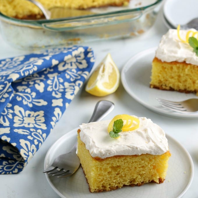slices of lemon cake garnished with mint leaves and lemon zest on white plates. In the background is the baking dish with the rest of the cake