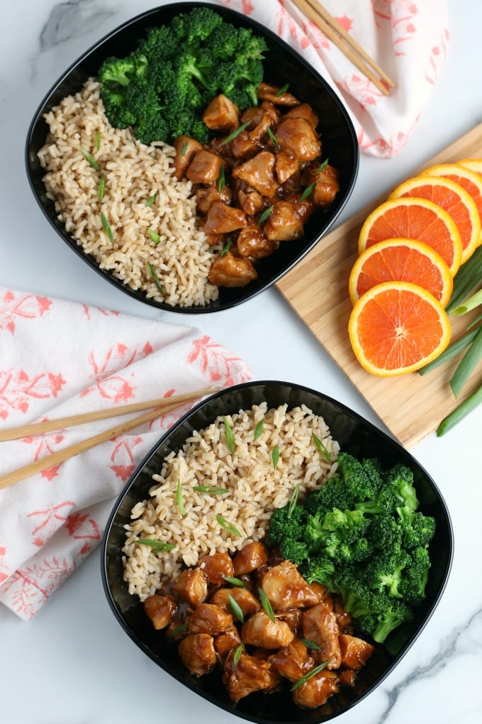 Top down image of instant pot orange chicken with rice and broccoli in a black bowl. Chopsticks and a napkin are off to the left. A cutting board with sliced oranges and green onions are on the right. 