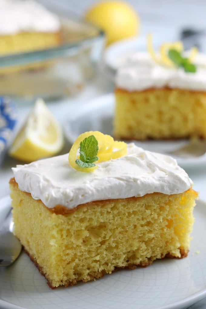 close up of a square piece of lemon cake with whipped topping "frosting," lemon zest and a tiny sprig of mint.
