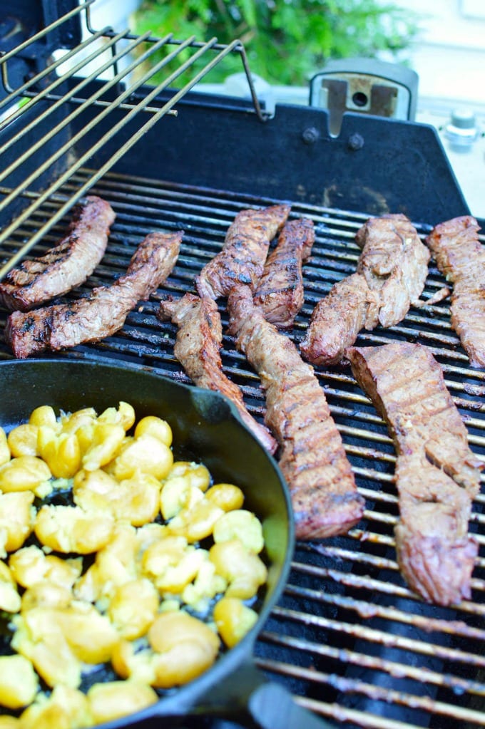 Grilled sirloin steak tips on a grill next to a cast iron skillet of potaotes.