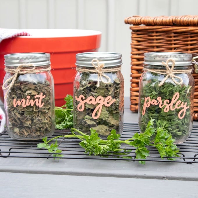 Three Cricut diy dried herb mason jars labeled with the words mint, sage, and parsley. Twine is wrapped around the neck of each jar. There is a large red bowl and a picnic basket in the background.