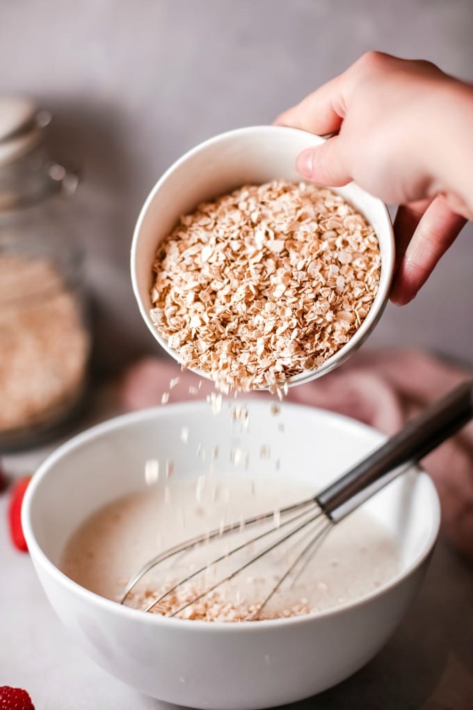Pouring oats into a large mixing bowl full of batter and a whisk.