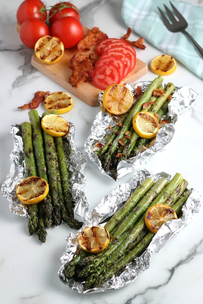 Easy Grilled Asparagus - foil pouches of grilled asparagus with grilled lemon. a butting board with tomatoes and bacon in the background.