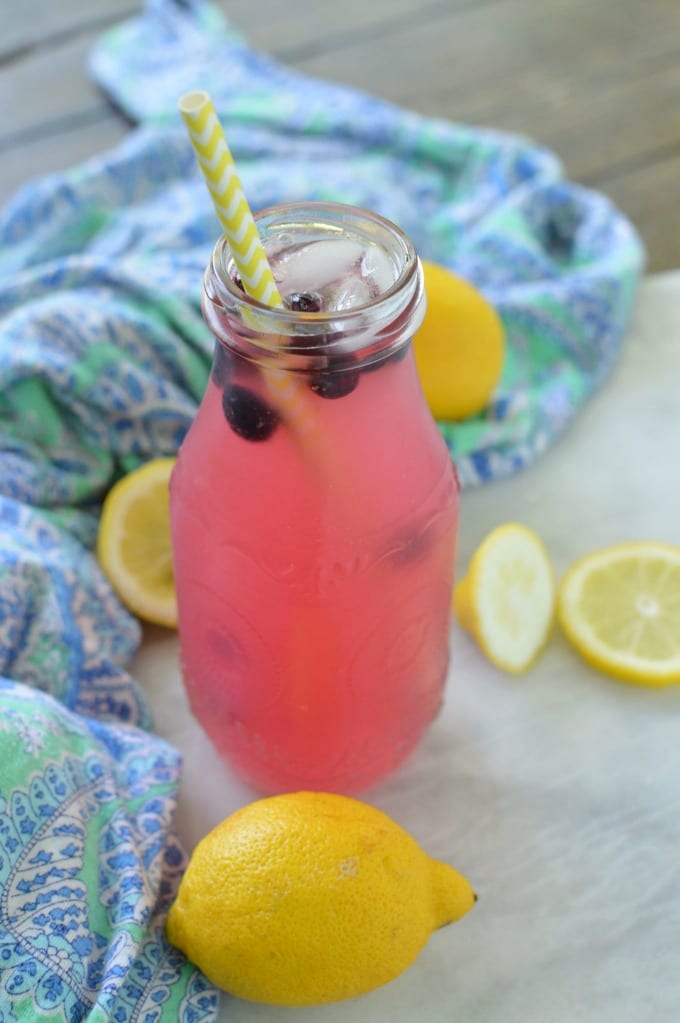 Blueberry lemonade recipe in a glass bottle with a paper straw, topped with fresh blueberries. The counter top is covered in lemon slices.