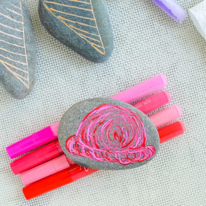 A smooth gray rock with a bright pink ice cream scoop painted on it laying on top of pink paint pens.
