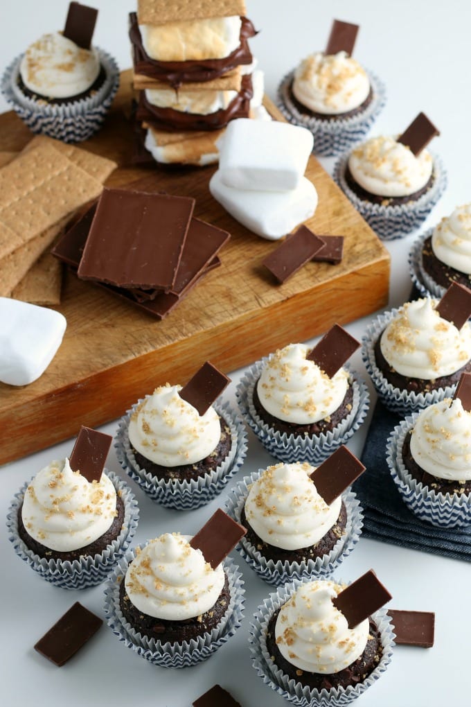 Chocolate cupcakes, topped with marshmallow creme frosting and chunks of chocolate, displayed with graham crackers, chocolate bars and marshmallows.