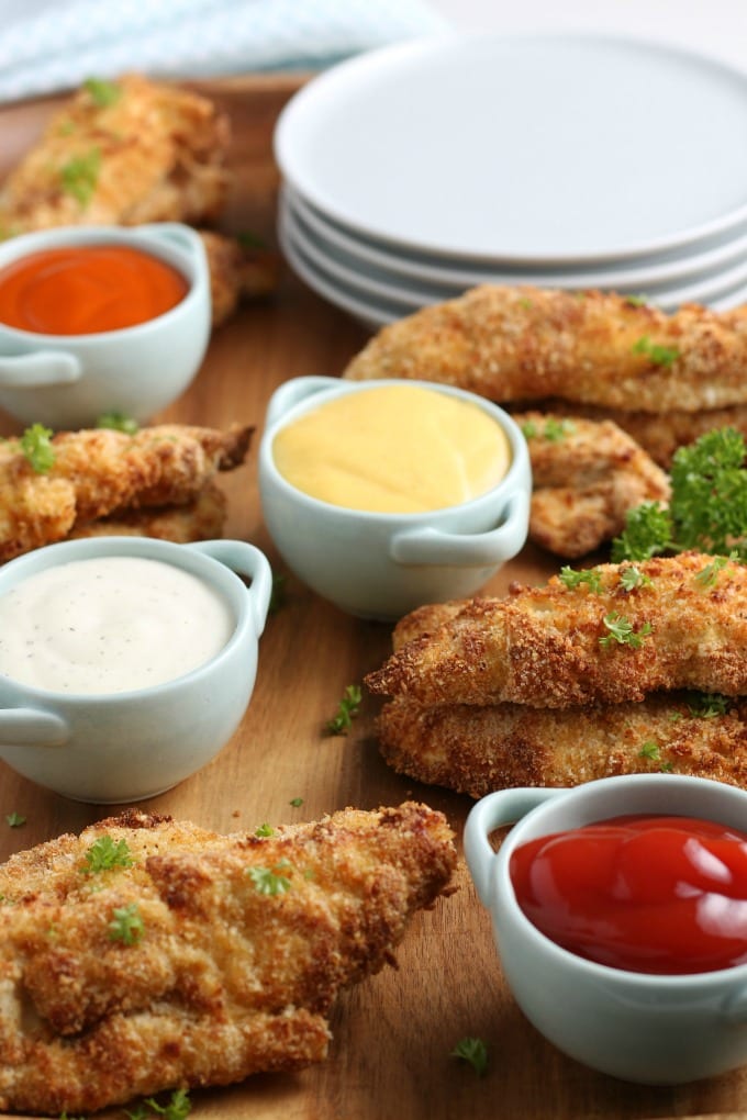 Golden brown air fried chicken tenders on a cutting board surrounded by ketchup, honey mustard and ranch sauces. 